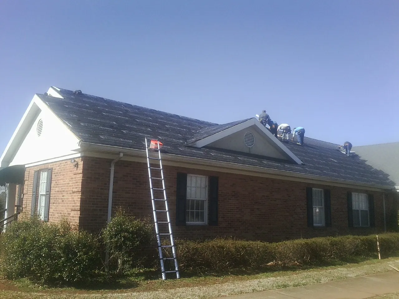 Crew tearing shingles off a brick ranch roof with a ladder set — Metal Roof Repair work by our crew in West Henrietta