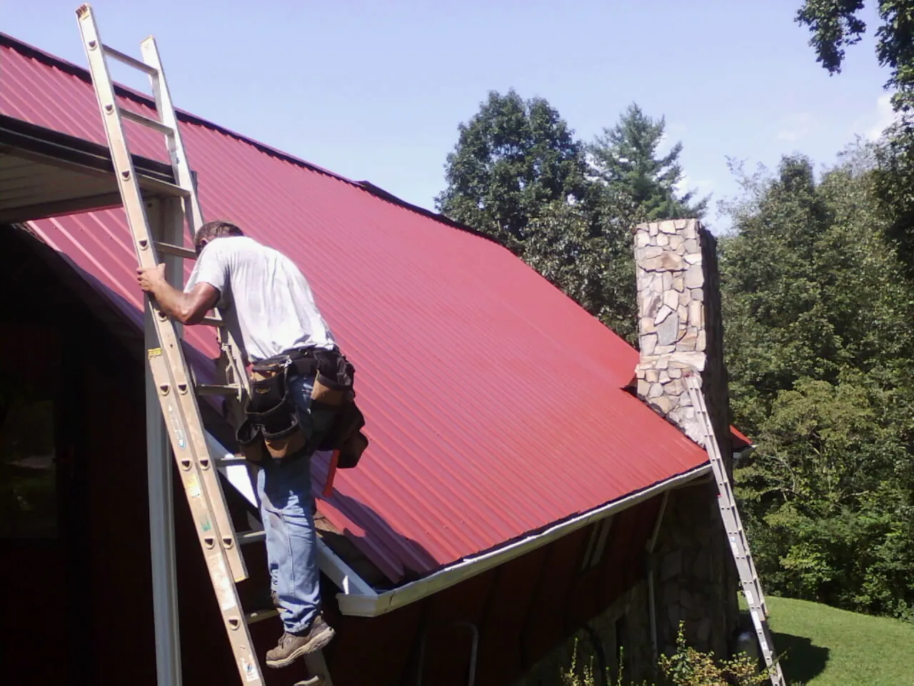 Roofer on a ladder beside a red metal roof and stone chimney — Soffit & Fascia Repair by Roofing Brothers in West Henrietta